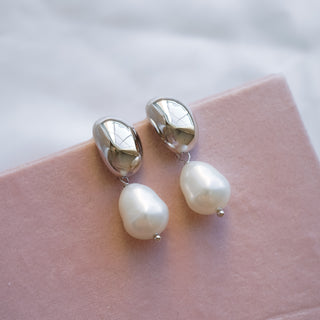 A pair of gold dome pearl drop earrings resting on a textured surface with a background of white satin.