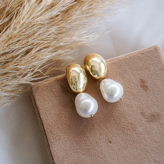 A pair of dome-shaped gold earrings with pearl drops displayed on a beige surface, with a fluffy beige plant in the background.