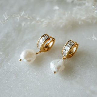 A pair of gold-colored hoop earrings with wire-wrapped pearls and cubic zirconia accents, displayed on a white fluffy surface.
