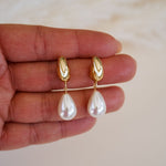 Gold earrings with pearl drops held between fingers on a light background