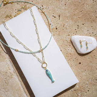 Necklace with fish pendant on a white card, earrings on a stone, on a beige surface