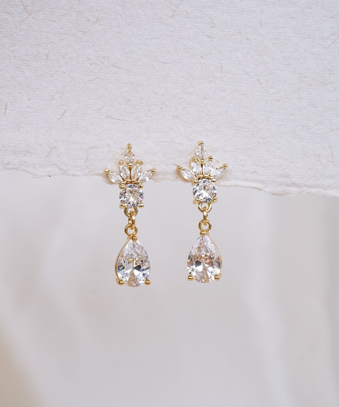 A pair of gold drop earrings with clear teardrop-shaped crystals, displayed against a soft white textured background.
