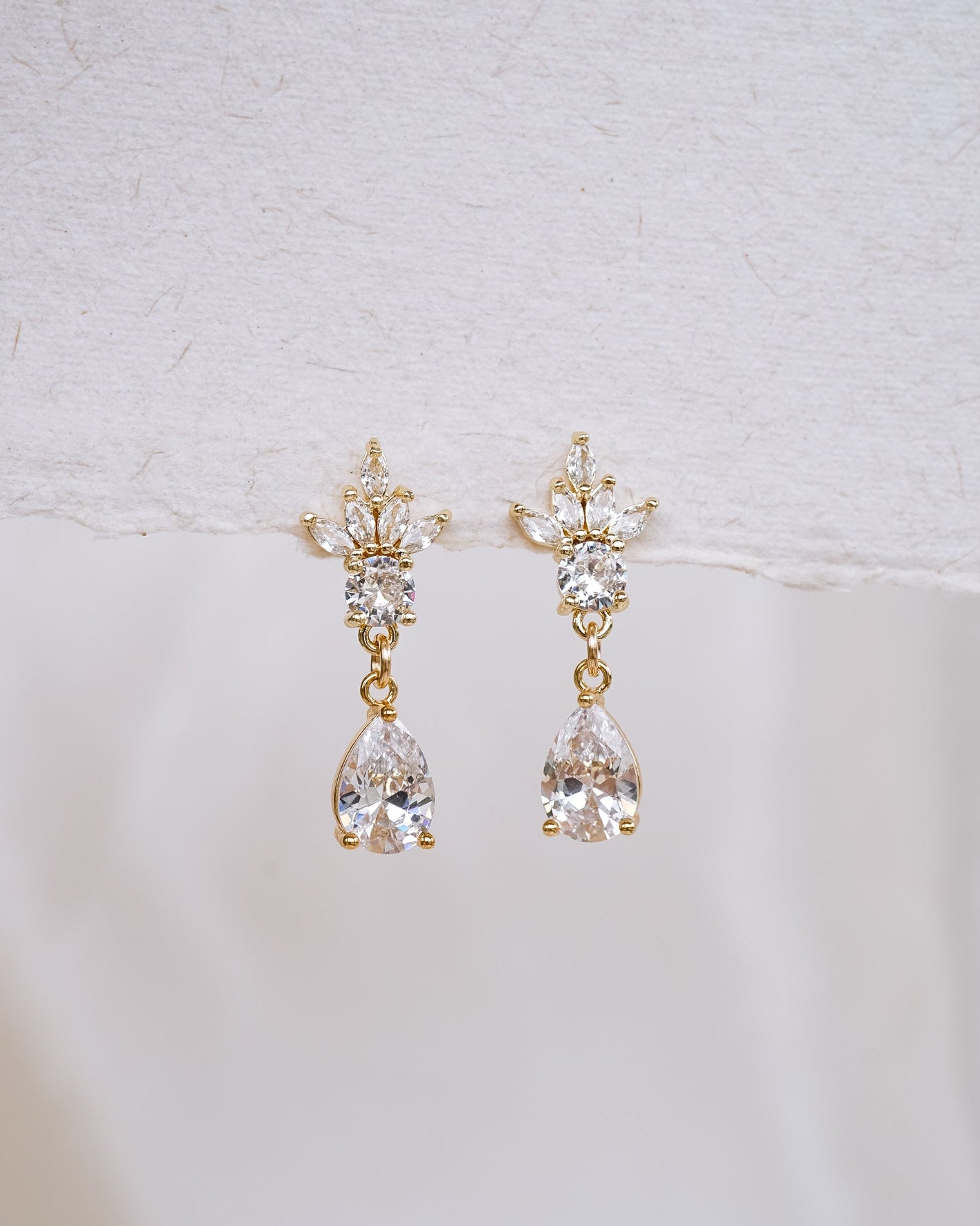 A pair of gold drop earrings with clear teardrop-shaped crystals, displayed against a soft white textured background.