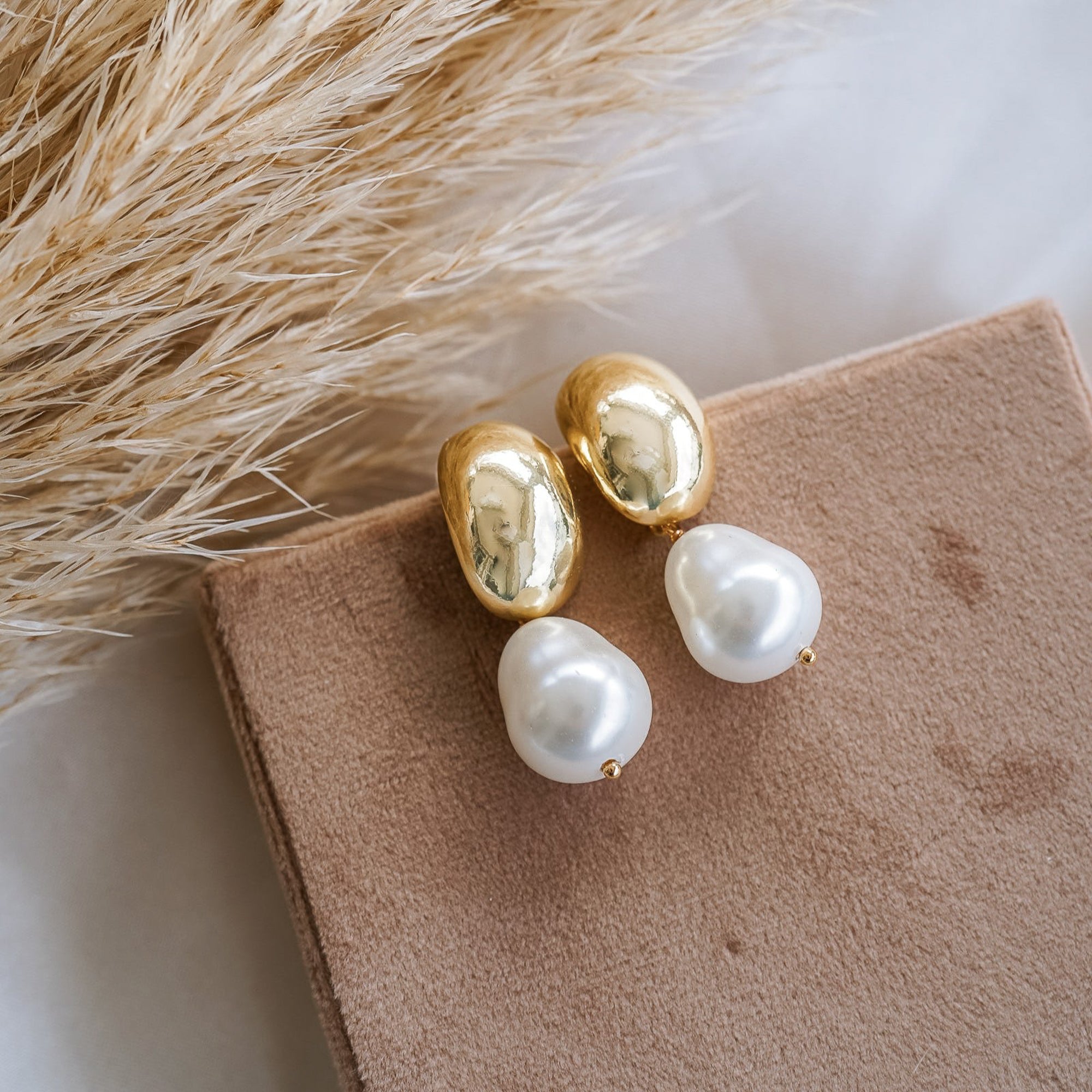 A pair of dome-shaped gold earrings with pearl drops displayed on a beige surface, with a fluffy beige plant in the background.