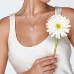 Woman wearing a gold necklace with a pearl pendant, holding a white flower against a plain background